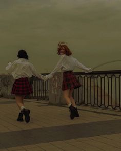 two young women dressed in plaid skirts are holding hands and walking on a bridge over looking the water