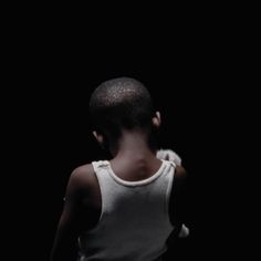 a young boy standing in the dark with his back turned to the camera, wearing a white tank top