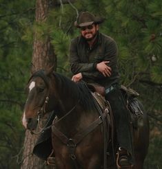 a man riding on the back of a brown horse in front of a forest filled with trees