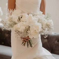 a bride holding a bouquet of white flowers in her wedding dress and standing next to a couch