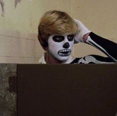 a man with white face paint sitting in front of a wall and holding his hands to his head