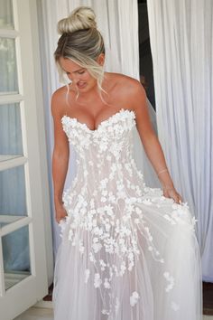 a woman in a white wedding dress is looking down at her dress while standing by the window