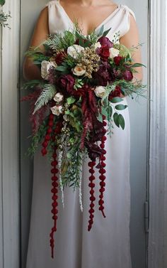 a woman in a white dress holding a red and green wedding bouquet with greenery