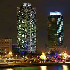 two tall buildings are lit up at night by the water in front of some lights