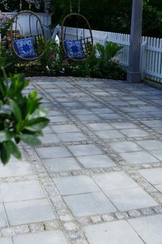 two hanging chairs on a brick patio in front of a white picket fence