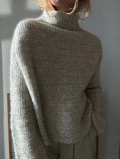 a woman wearing a turtle neck sweater and pants in front of a white wall with sunlight coming through the window