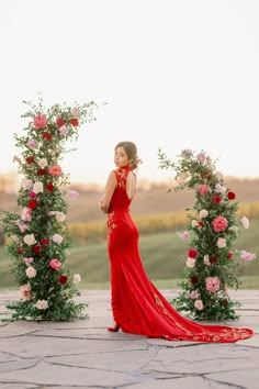 a woman in a long red dress standing next to flowers and greenery with her hand on her hip