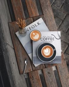 two cups of coffee sitting on top of a wooden table next to an open book