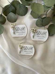 three white plates with flowers and names on them sitting next to some green leaves in the background