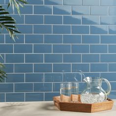 two glasses and a pitcher on a tray in front of a blue tiled wall with palm leaves