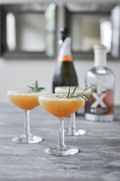 two wine glasses filled with orange juice and garnished with rosemary sitting on a table