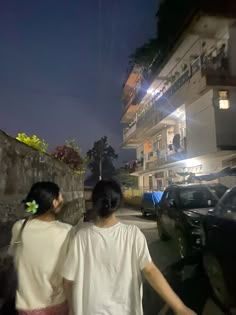 two people walking down the street at night with cars parked in front of them and an apartment building behind them