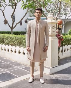 a man standing in front of a white fence wearing a beige outfit and shoes with his hands on his hips