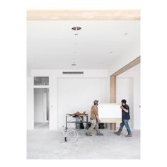 two men are standing in an unfinished room with white walls and floors, one man is painting the wall