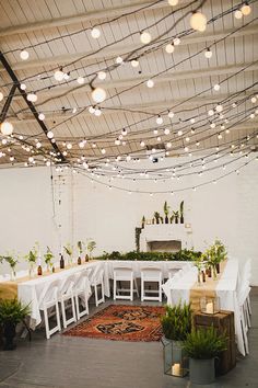 a room with tables and chairs covered in white tablecloths, hanging lights and potted plants