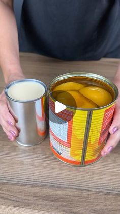 a woman holding two cans of paint on top of a wooden table next to a cup