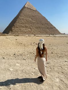 a woman walking in front of the giza pyramid, with a white hat on her head