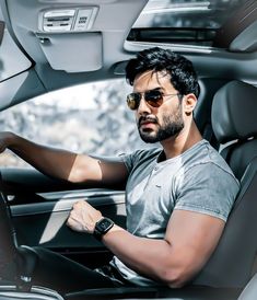 a man wearing sunglasses sitting in the driver's seat of a car with his hand on the steering wheel