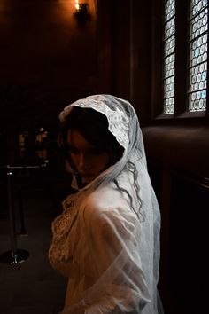 a woman in a white veil standing next to a window with stained glass behind her