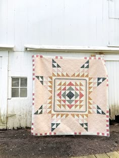 a quilt hanging on the side of a white building with a barn in the background