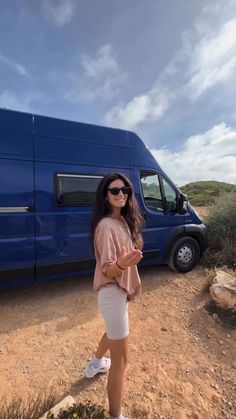 a woman standing in front of a blue van on the side of a dirt road