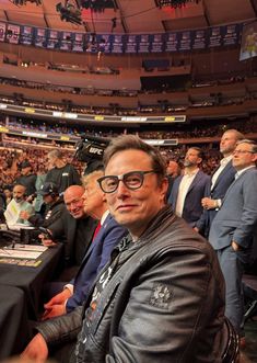 a man sitting at a table in front of a crowd wearing glasses and a leather jacket