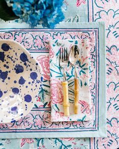 a place setting with blue and pink flowers on the table cloth, utensils and napkins