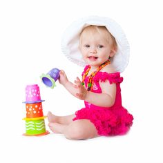 a baby girl in a pink dress and hat playing with a stack of colorful toys