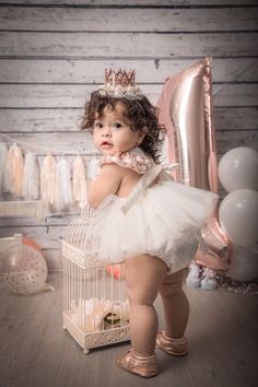 a baby girl in a white tutu and tiara standing next to a birdcage