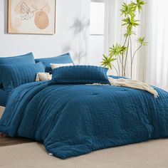 a bed with blue comforters and pillows in a room next to a plant on the floor