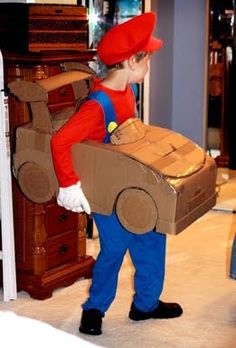 a young boy dressed in mario costume carrying a cardboard box with a car on it