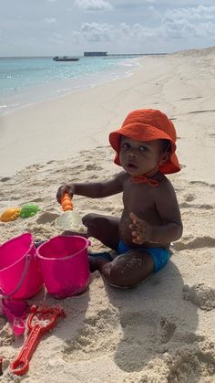 a baby sitting on the beach playing with buckets and sand while wearing an orange hat