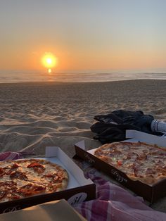 two pizzas sitting on top of boxes in the sand near the ocean at sunset