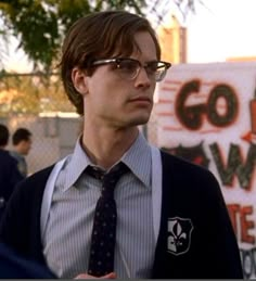 a man wearing glasses and a tie standing in front of a sign with writing on it