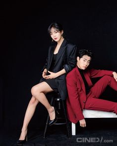 two people sitting on a chair in front of a black background, one wearing a red suit and the other a black shirt