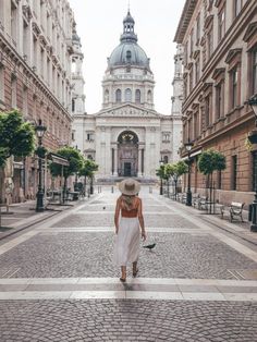 a woman in a white dress and straw hat walking down an empty street with buildings on both sides