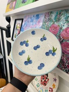 a person holding up a bowl with blueberries on it in front of some paintings