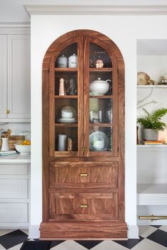 a wooden china cabinet with glass doors in a white kitchen, next to a black and white checkered floor