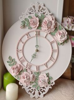 a white clock decorated with pink flowers and green leaves on a table next to candles