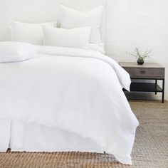 a bed with white sheets and pillows in a room next to a nightstand on the floor