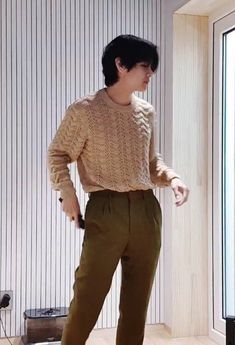 a young man standing in front of a window wearing brown pants and a beige sweater