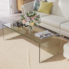 a glass coffee table with flowers on it in front of a white couch and chair