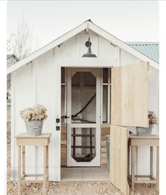 a white shed with two wooden tables in front of it and a potted plant next to the door