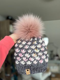 a person holding up a knitted hat with pink fur pom - pom