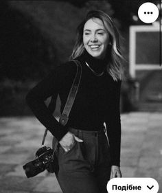 a black and white photo of a woman holding a camera smiling at the camera with her hand on her hip