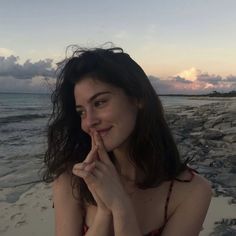 a woman sitting on top of a beach next to the ocean with her finger in her mouth