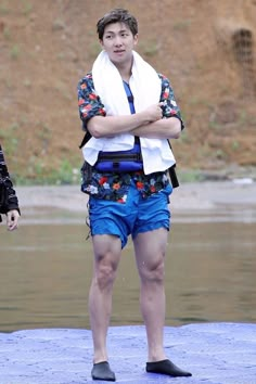 a man with his arms crossed standing in front of a body of water wearing blue shorts and a white towel