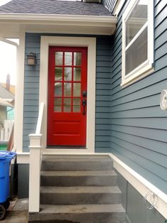 a red door on a blue house with steps leading up to the front door and trash can