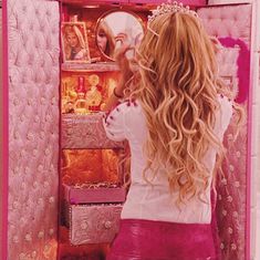 a woman with long blonde hair looking into a mirror in front of a pink wall