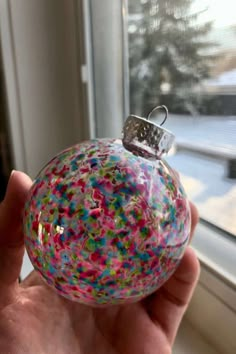 a hand holding a glass ornament with sprinkles on it in front of a window